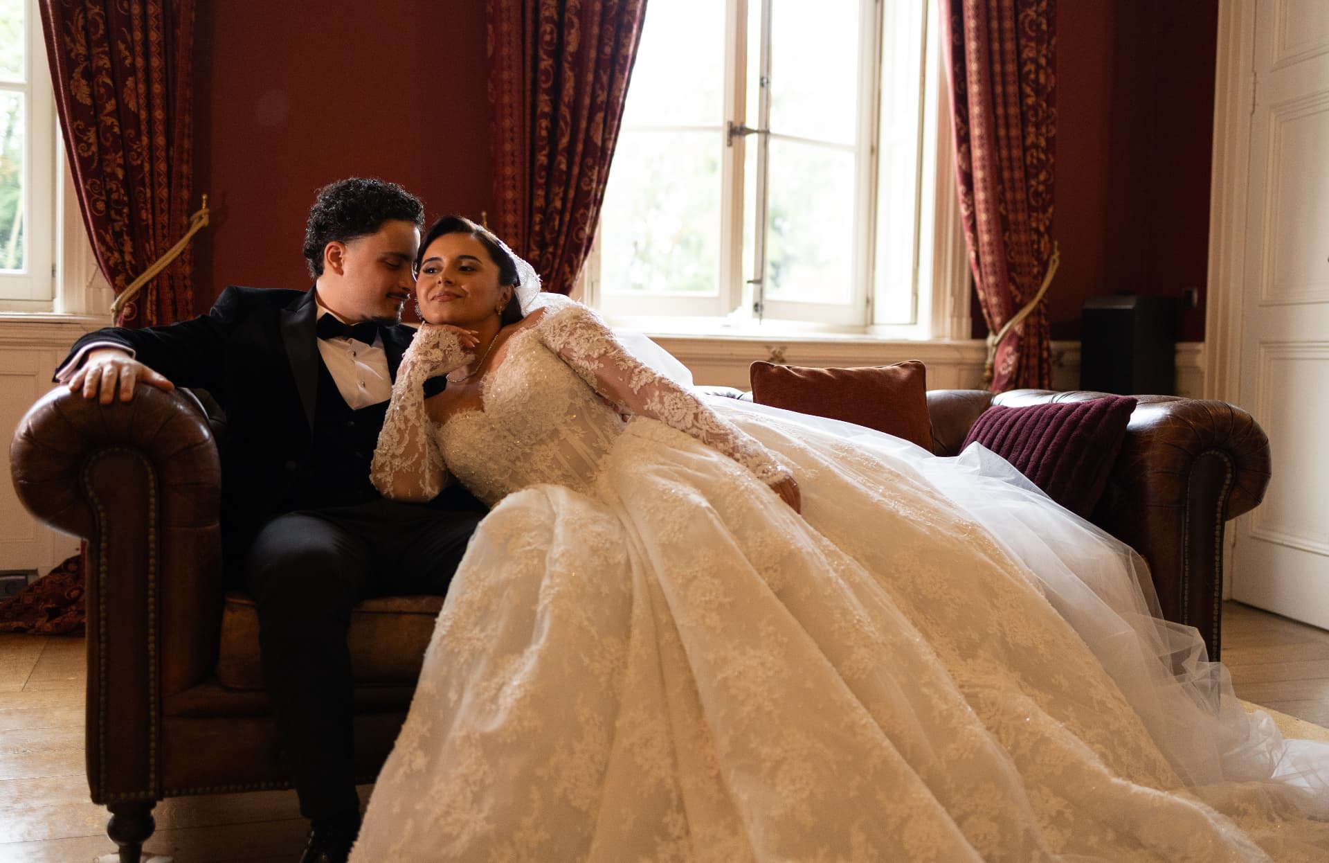 Newlyweds seated together on a sofa beside tall windows