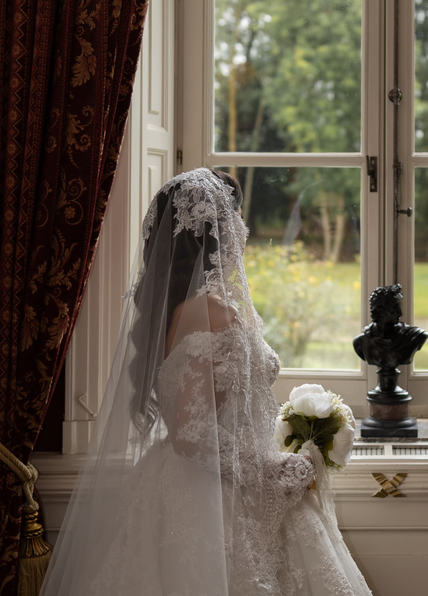A bride holding a white bouquet beside a window overlooking greenery