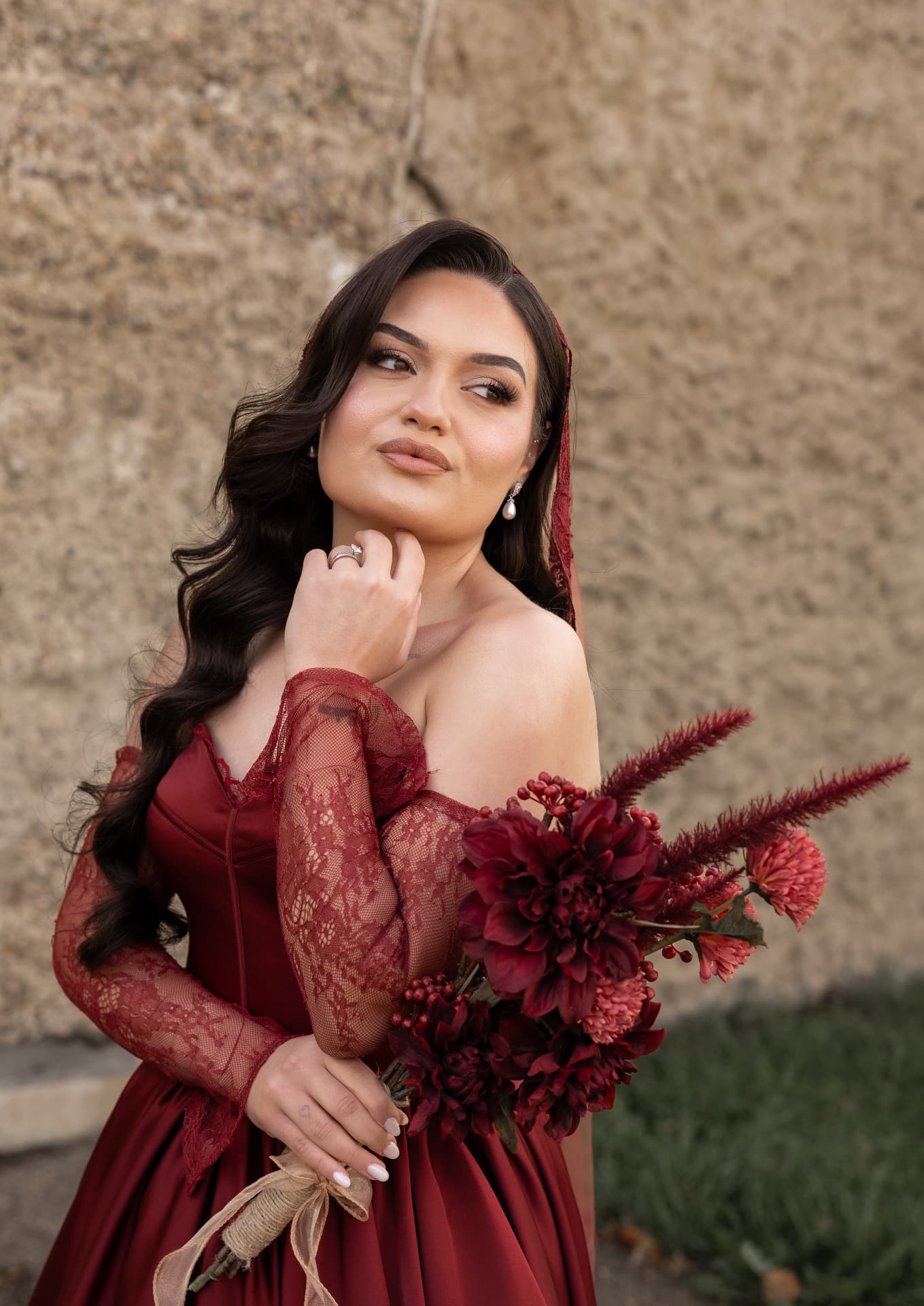 A bride in a burgundy satin gown with lace sleeves holding a deep red bouquet